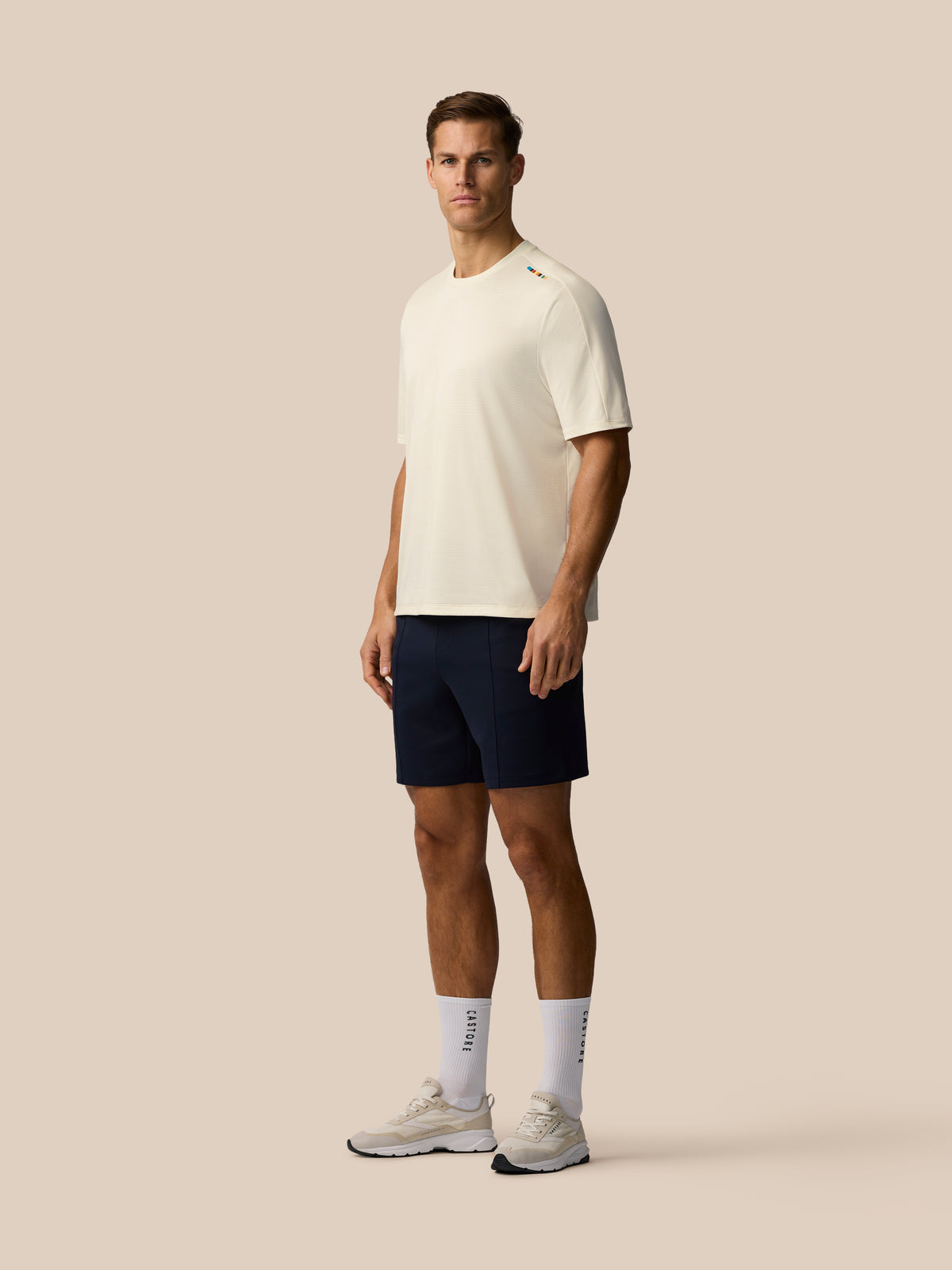 Full body view of a man in a cream knit T shirt navy shorts and sneakers against a beige background