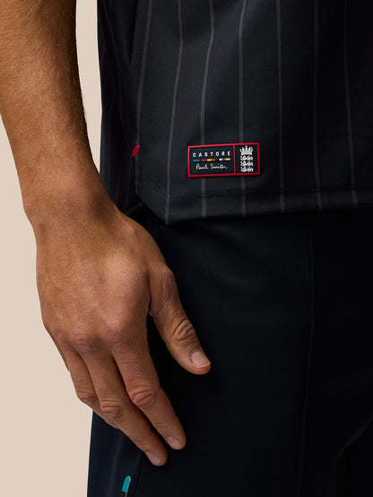 Close-up of the shirt shows a colorful Castore Paul Smith patch at the lower front hem on a dark striped T-shirt.