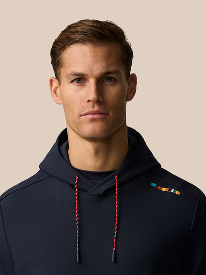 Full body view of a man in a navy hoodie with red drawcords and a small multicolor signature stripe on the left shoulder