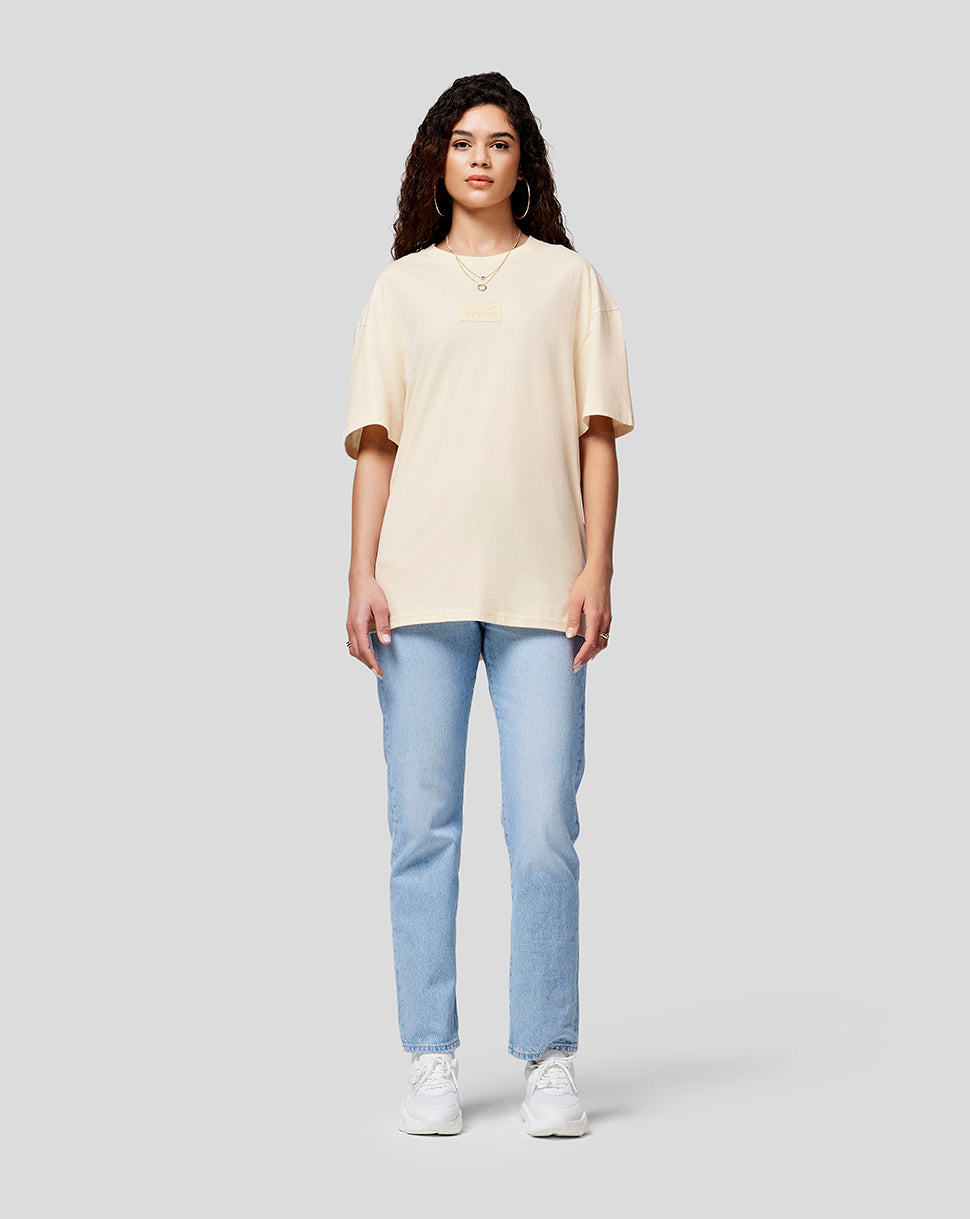 Front view of a woman wearing a cream oversized T shirt with chest patch, light wash jeans and white sneakers.