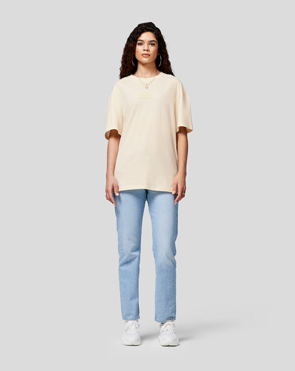 Front view of a woman wearing a cream oversized T shirt with chest patch, light wash jeans and white sneakers.
