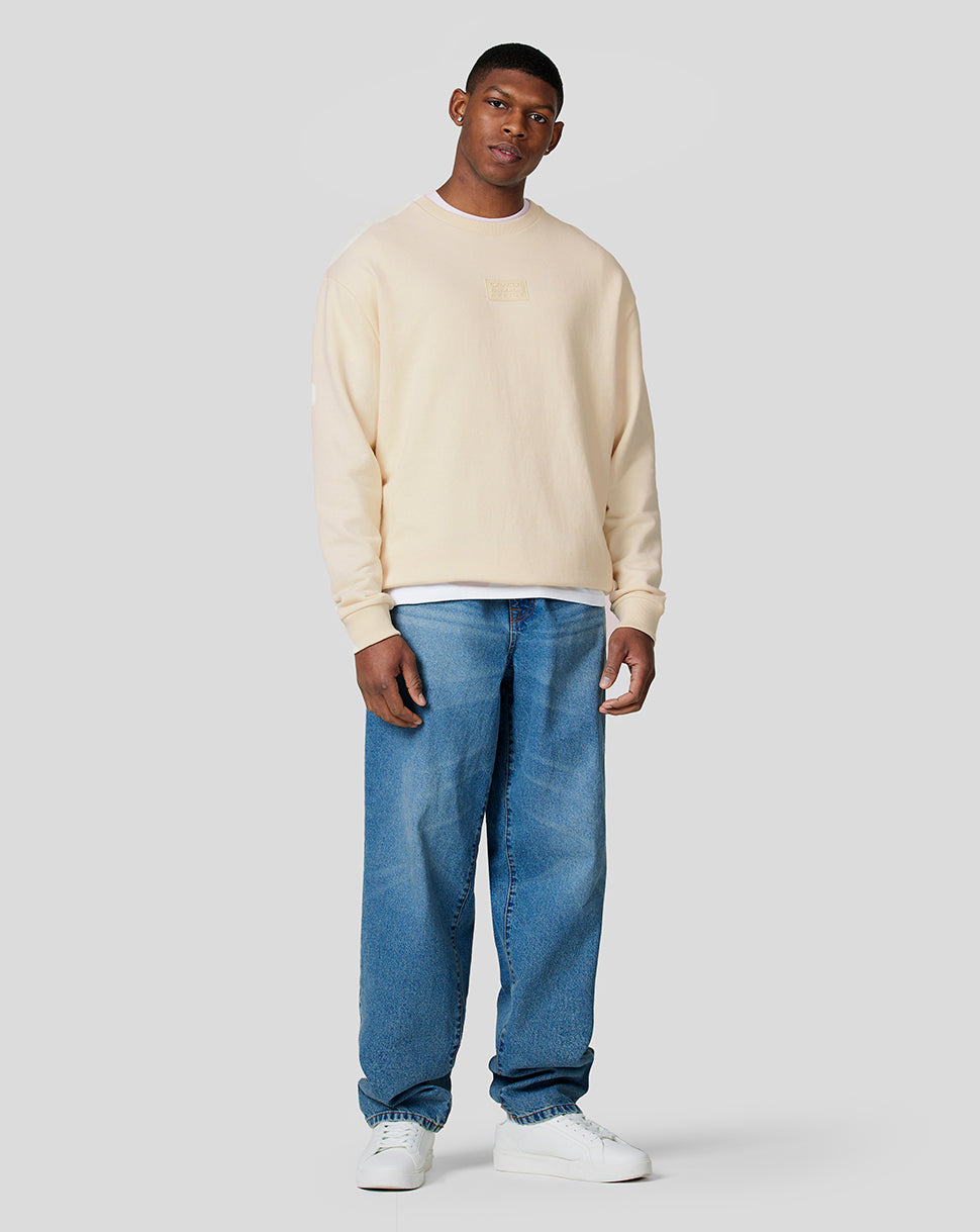 Full-body view of a male model in an ecru oversized crew neck sweatshirt with chest patch and white tee underneath