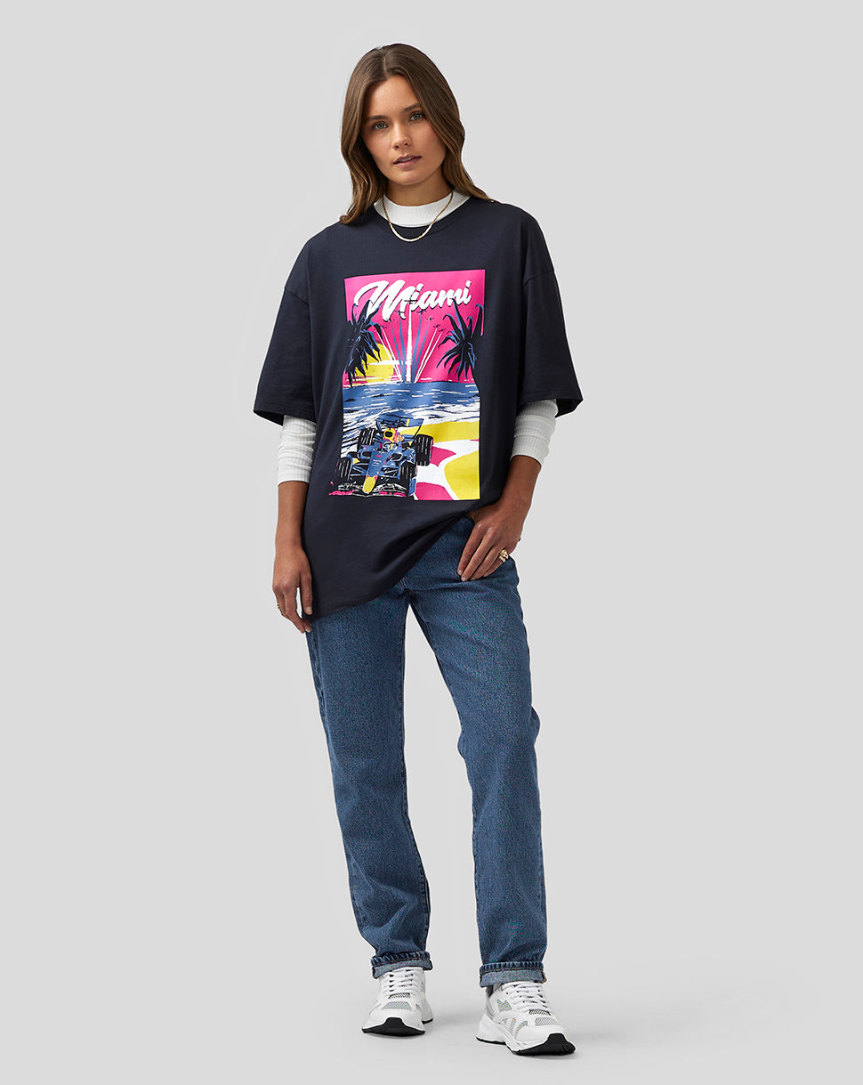 Full body of a female model wearing a navy Miami graphic oversized T shirt over a white long sleeve with jeans.