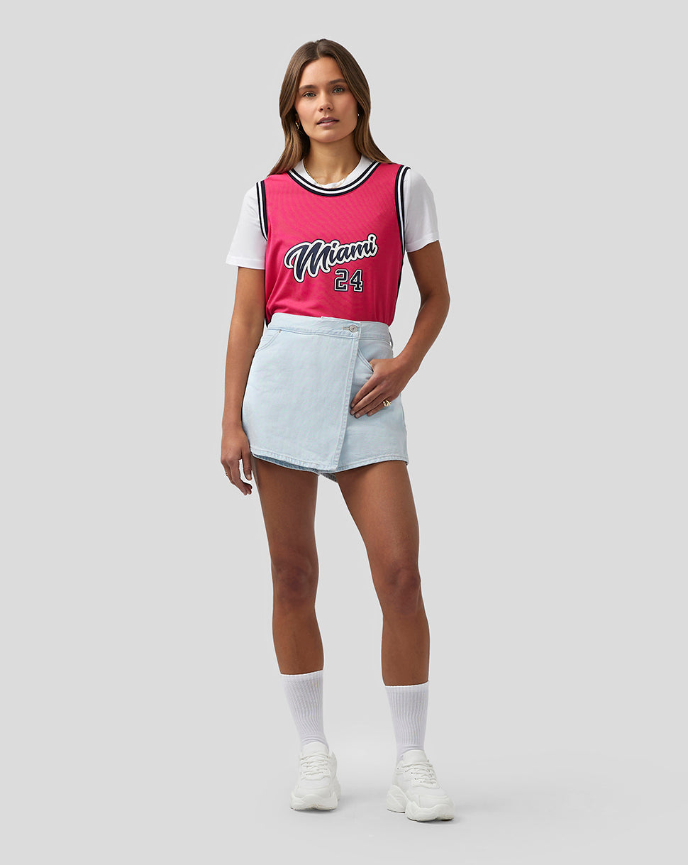 Full body shot of a model wearing a bright pink Miami basketball jersey with white tee underneath and denim skirt