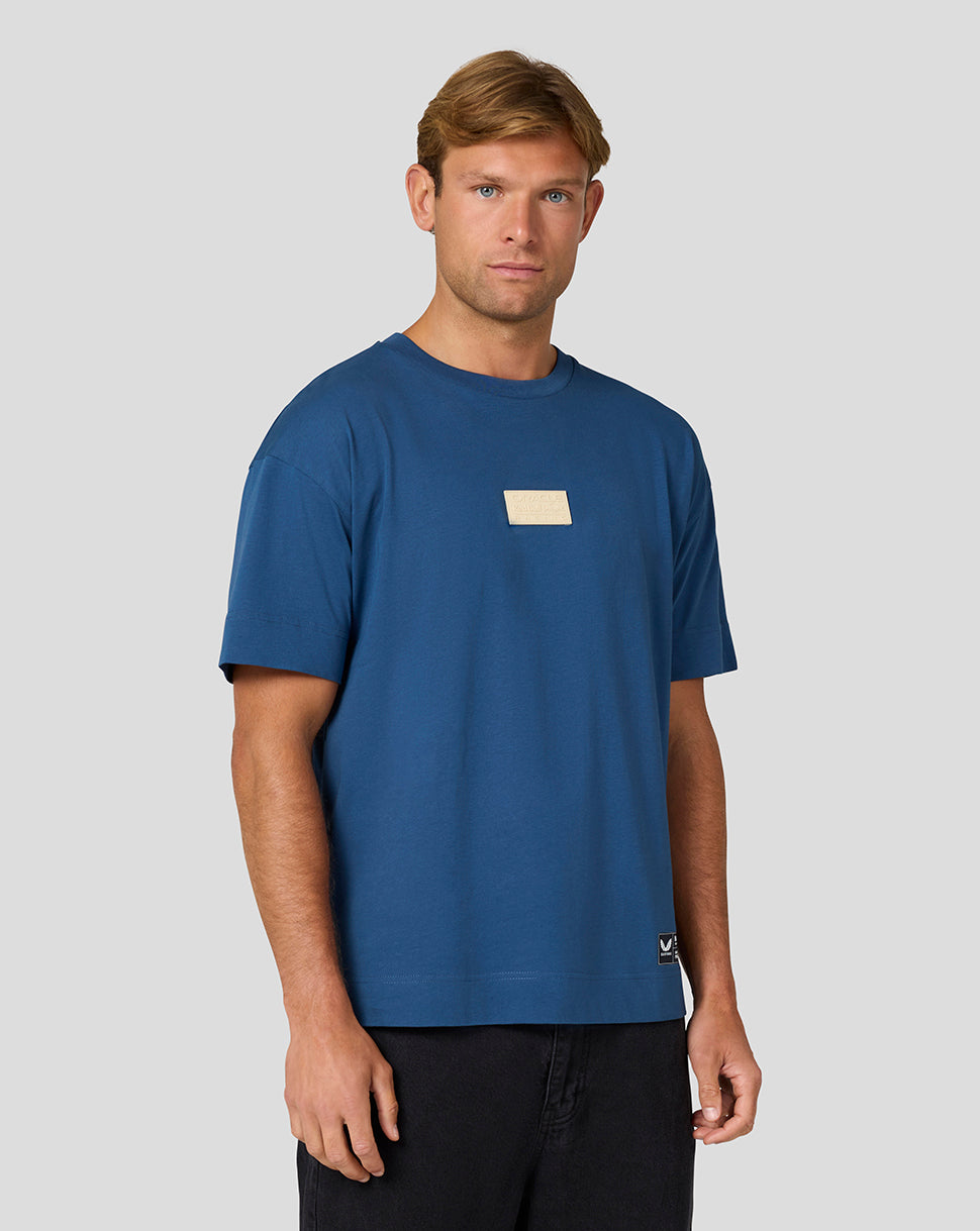 Male model wearing blue short sleeve tee with beige plaque logo on chest.