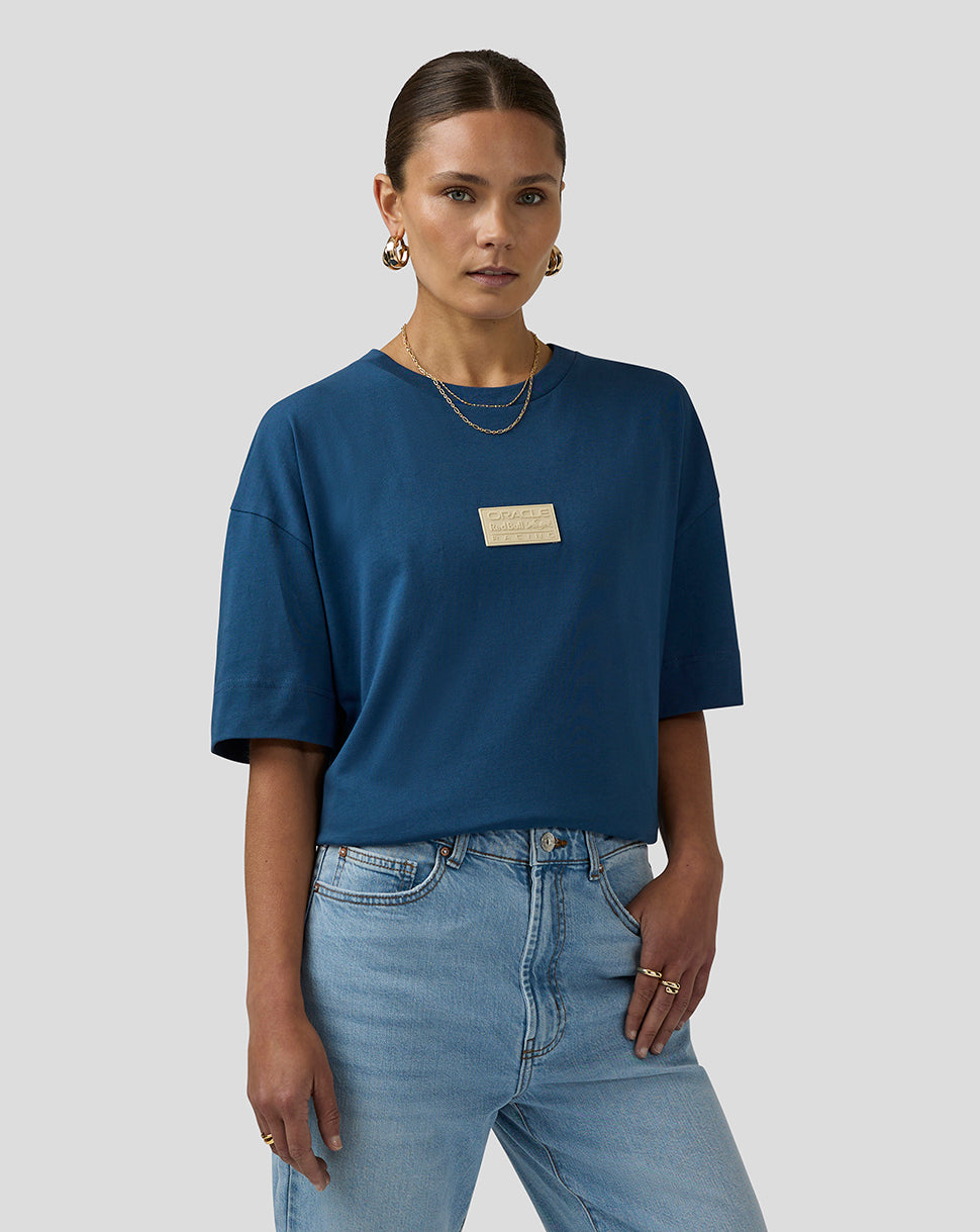 Female model wearing blue short sleeve tee with beige chest plaque and light wash jeans.