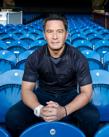 Man wearing black Rangers Retro 2002-03 Home Blackout shirt seated in blue stadium seats