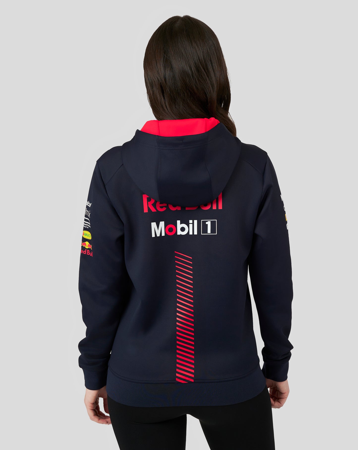 Female model in a navy Red Bull Racing full zip hoodie seen from the back with red interior and a large Red Bull logo; front pocket logos visible.
