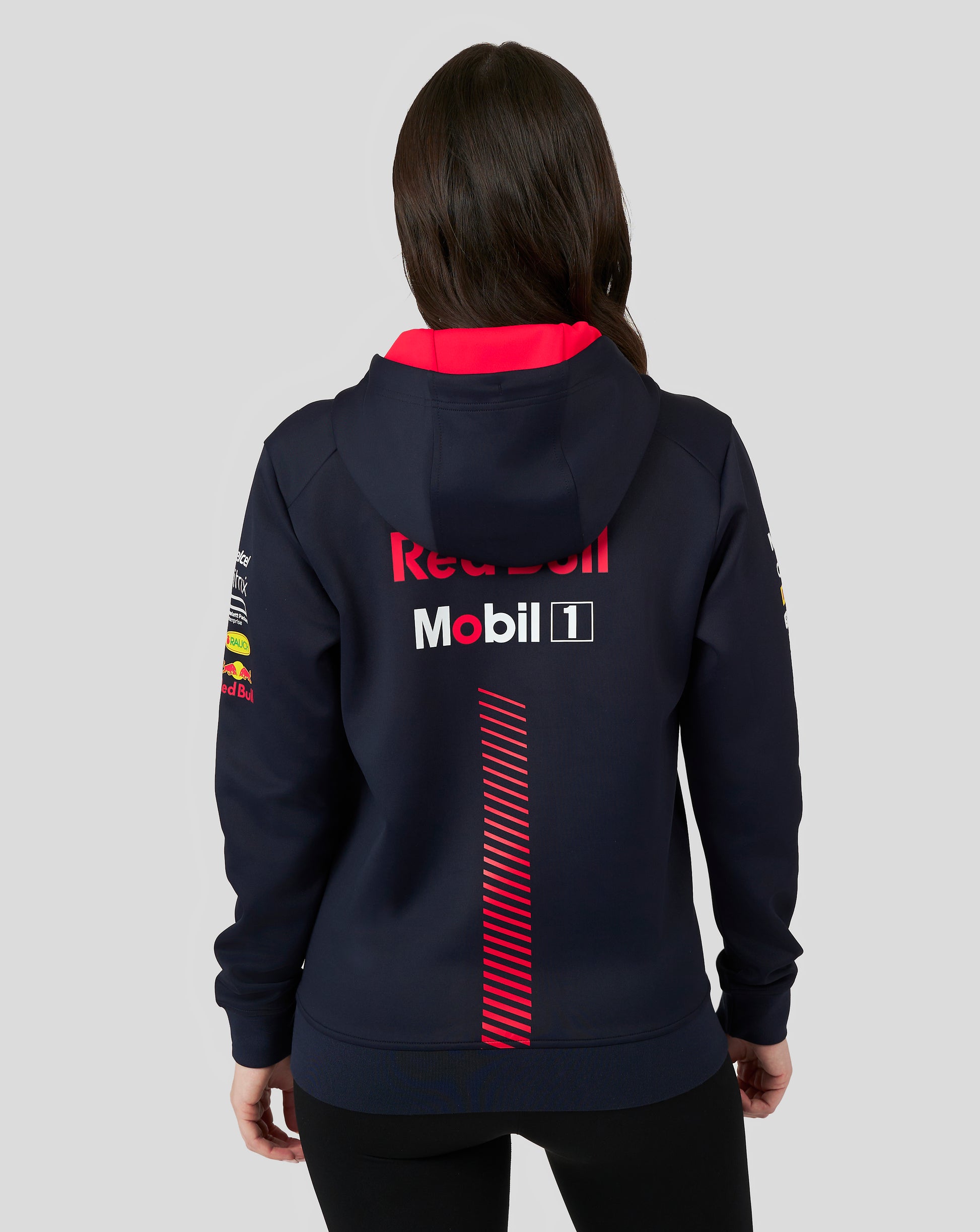 Female model in a navy Red Bull Racing full zip hoodie seen from the back with red interior and a large Red Bull logo; front pocket logos visible.