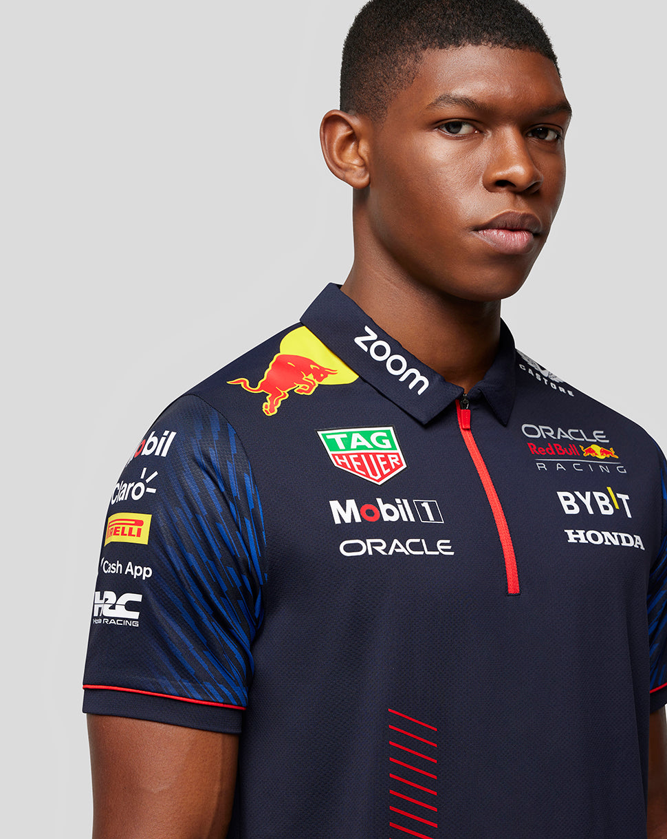 Male model in navy polo with numerous sponsor logos on the chest and a red zip placket.