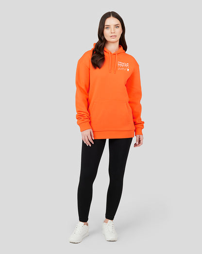 Full-body shot of a woman in an orange Oracle Red Bull Racing hoodie paired with black leggings and white sneakers.
